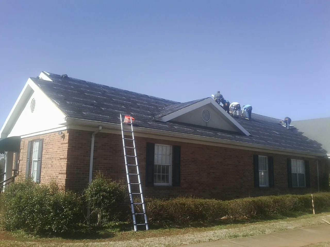 Crew tearing shingles off a brick ranch roof with a ladder set — Emergency Roof Repair work by our crew in Plumtree