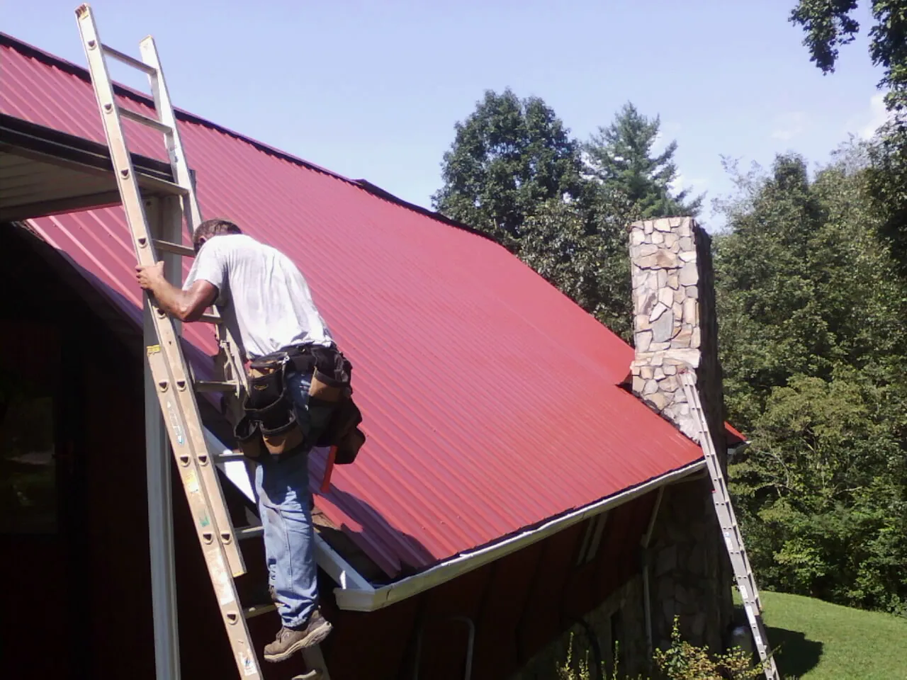 Roofer on a ladder beside a red metal roof and stone chimney — Soffit & Fascia Repair by Roofing Brothers in Plumtree